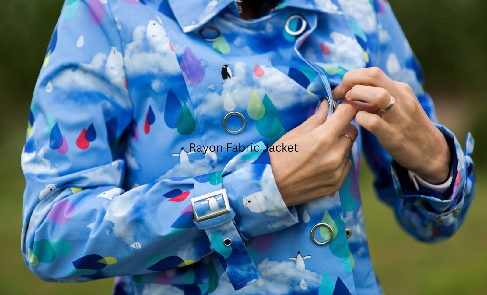 Rayon Fabric Jacket: Perfect Blend of Style & Softness “Close-up of a light blue rayon fabric jacket with a colorful, weather-themed print featuring clouds, raindrops, and small shapes. A person is fastening the jacket near the chest area; text on the jacket reads ‘Rayon Fabric Jacket.