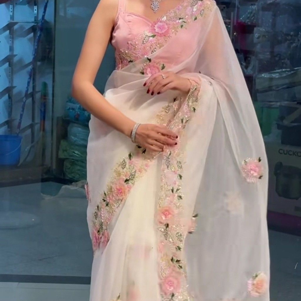 A woman wearing a sheer, pale pink saree with delicate pink floral embroidery draped over a pink blouse. She stands indoors, posing with one hand resting on the fabric and the other lightly touching the edge of the saree; subtle jewelry and red nail polish complete the look.