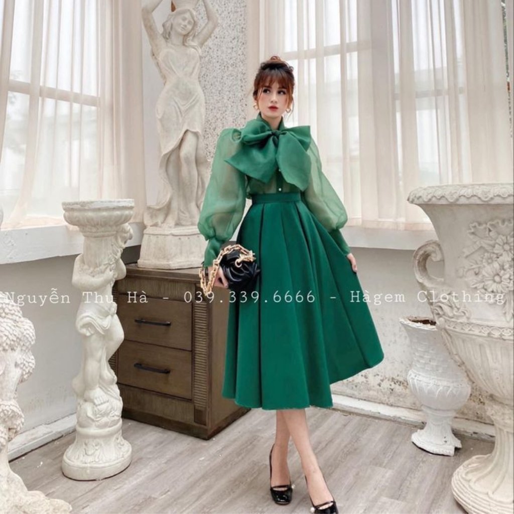 A stylish woman stands indoors in a green satin organza dress with a voluminous puff-sleeve bodice and a large bow at the neckline. She pairs it with black high heels and a small handbag, posing among vintage white sculptures and ornate planters.