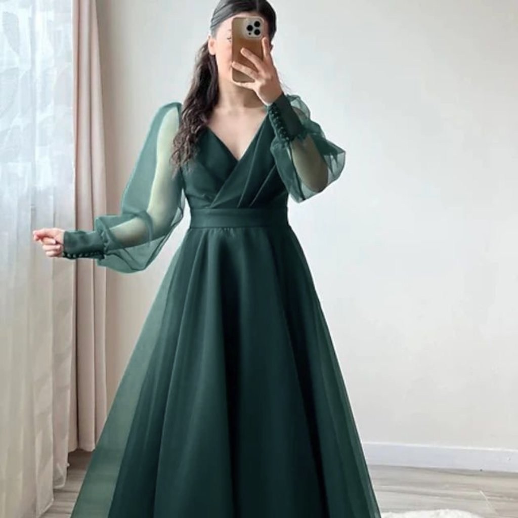 A full-length emerald-green wedding dress with a fitted bodice and a flowing satin organza skirt. The dress features sheer, puffed long sleeves and a V-neckline created by draped satin overlays. The wearer holds a phone in front of their face, standing in a softly lit room with pale walls and sheer curtains.