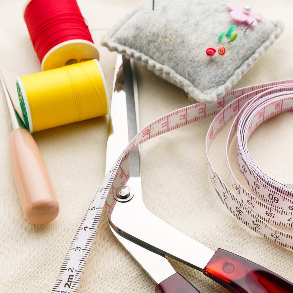 How to Sew Organza Fabric Like a Pro: Essential Sewing Tips 11 A close-up of sewing supplies: red and yellow spools of thread, a beige fabric pin cushion with pins, a measuring tape, a pair of silver scissors, and a brown seam-ripper or tool handle on a light background.