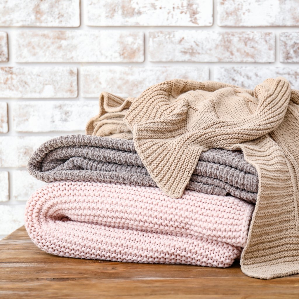 A stack of folded knit blankets in soft neutrals (pink, gray, and beige) with a beige throw draped over the top, set on a wooden surface against a white brick wall.