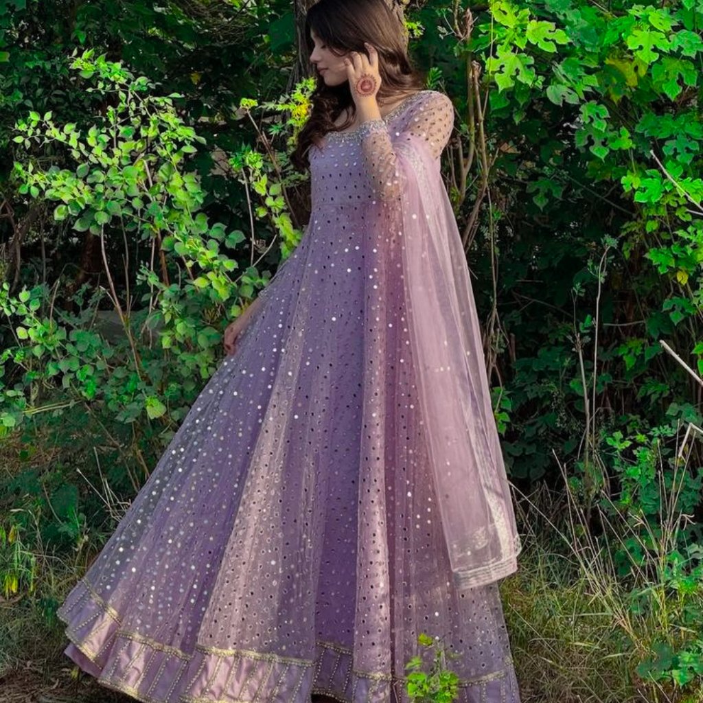 A woman wearing a lavender-to-pink gradient, full-length, sparkling organza Anarkali dress with dotted embellishments and a sheer cape, standing in a lush green garden.