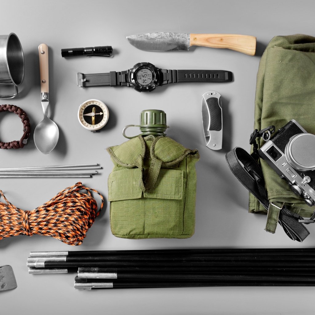 A flat lay of outdoor survival gear spread on a gray surface, including a green tactical pouch, a green canteen cover, a black digital watch, a knife with a wooden handle, a compass, a folding knife, a camera with strap, a coil of orange paracord, metal stakes, tent poles, a metal mug, and various hiking accessories.