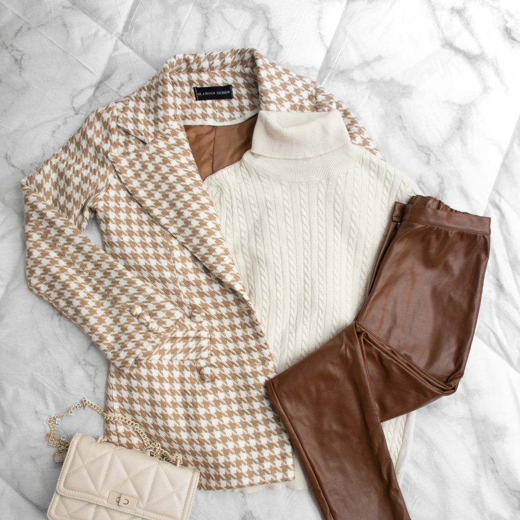 An on-trend outfit laid flat on a light marble surface: a beige-and-brown houndstooth blazer, a cream cable-knit turtleneck, glossy brown leather pants, and a small cream quilted handbag with a chain strap.