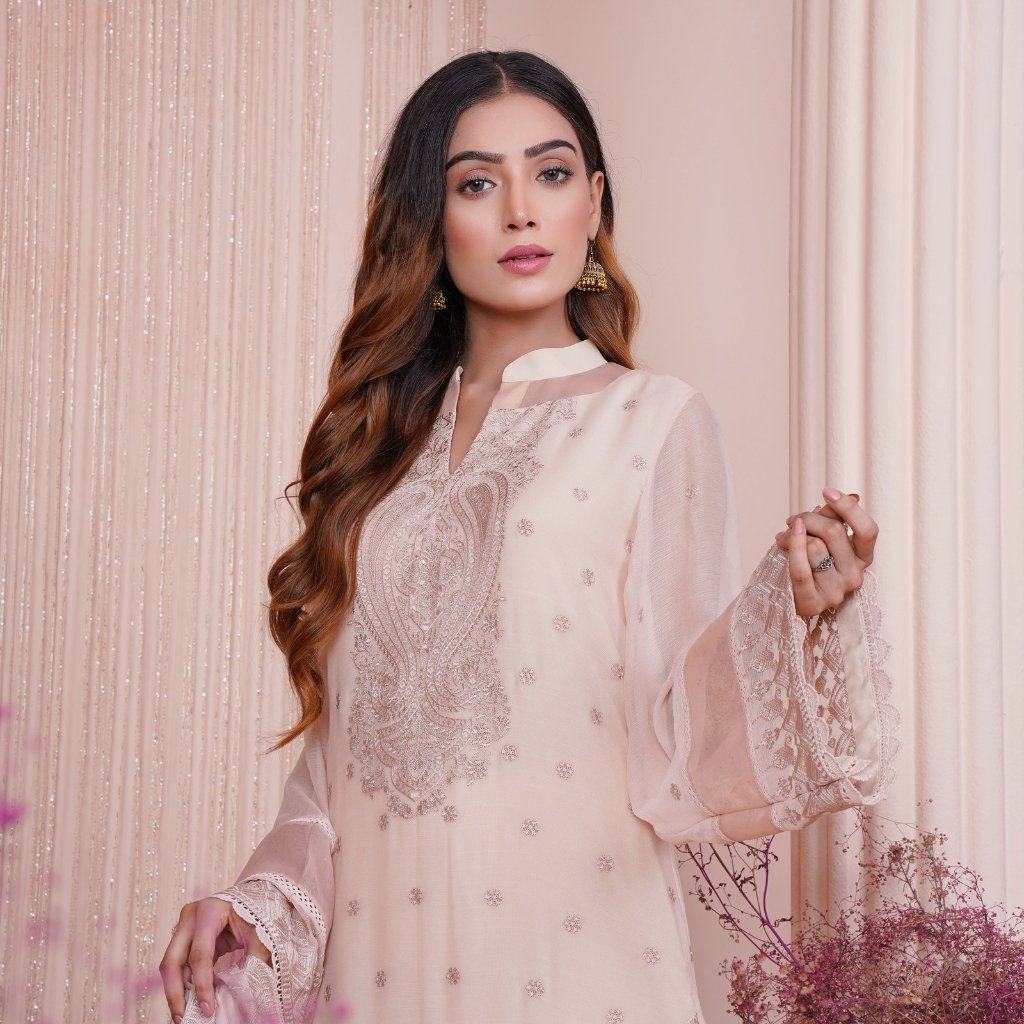 A woman wearing a light pink embroidered kurta with intricate silver threadwork and sheer sleeves, standing against a soft pink backdrop.