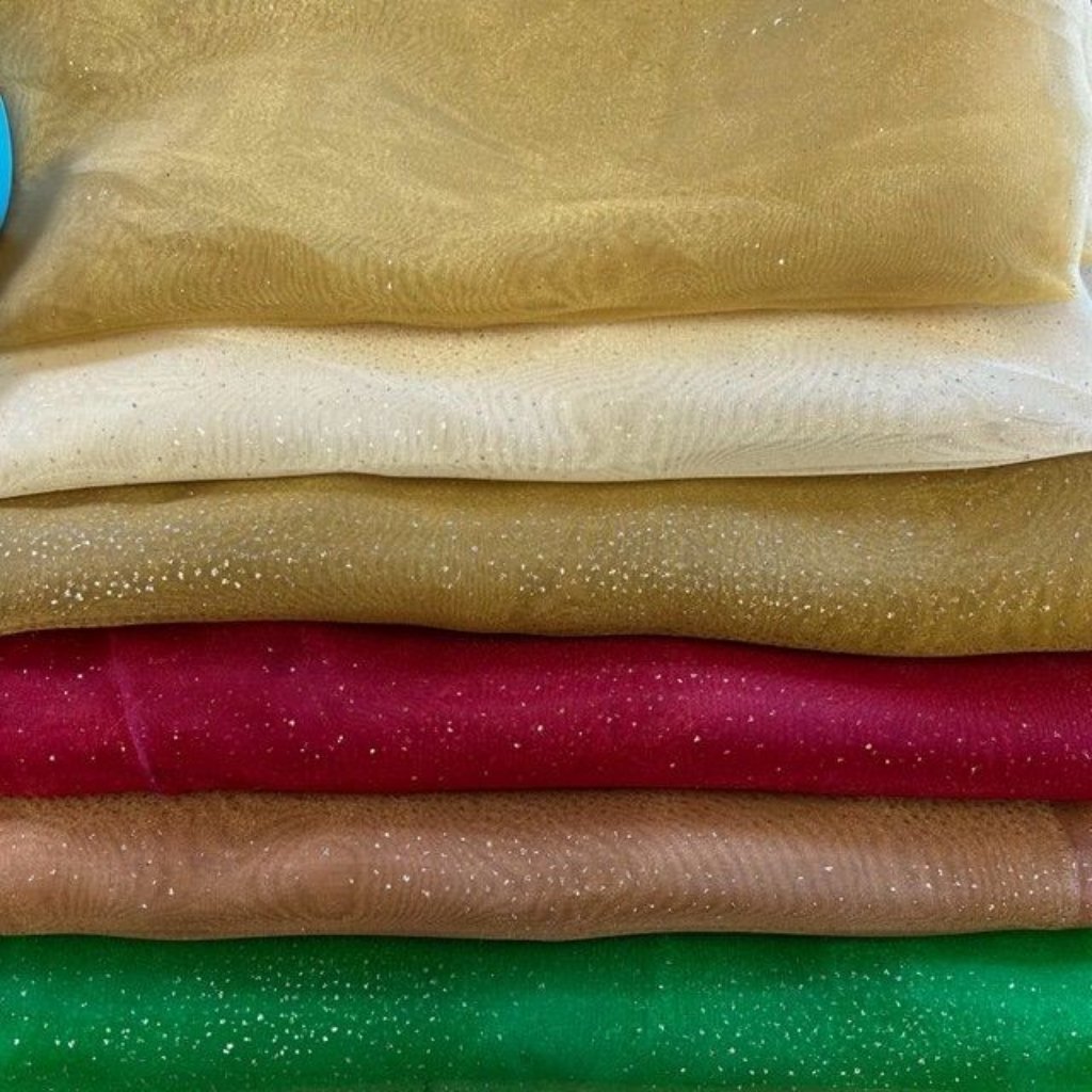 Stacked folded fabrics in a rainbow of colors from top to bottom: gold, cream, tan, magenta, light brown, and green, with small white specks scattered across the surfaces.