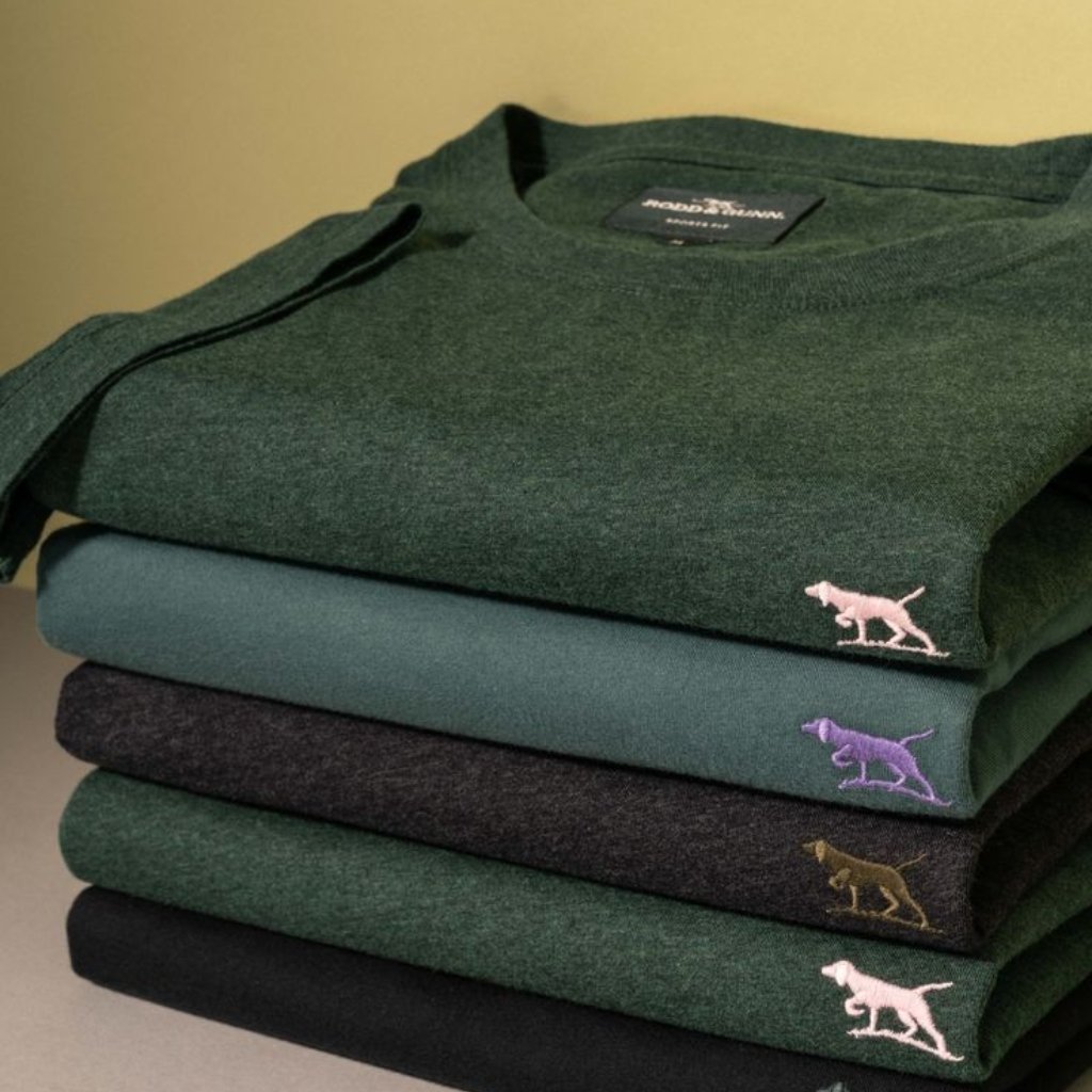 A neatly stacked pile of folded crewneck sweaters in earthy tones (green, teal, charcoal, and black). Each sweater features a small embroidered collared dog logo near the bottom edge; the top green sweater shows a pink dog, the next teal shows a purple dog, the third charcoal shows a brown dog, and the bottom black shows a pink dog. A Rodd & Gunn label is visible on the top sweater.