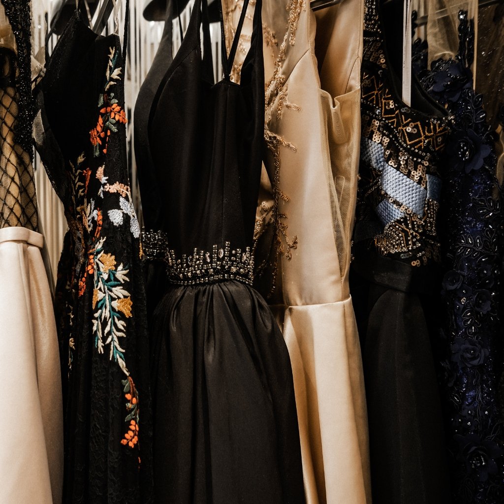 A row of elegant evening gowns in black, cream, and navy, featuring embroidered florals, sequin accents, and satin textures, hung on a wardrobe rack.