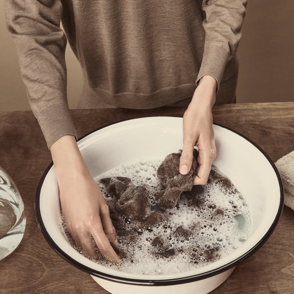 A person wearing a brown sweater washes multiple dark fabric pieces in a white enamel bowl filled with soapy water on a wooden table.