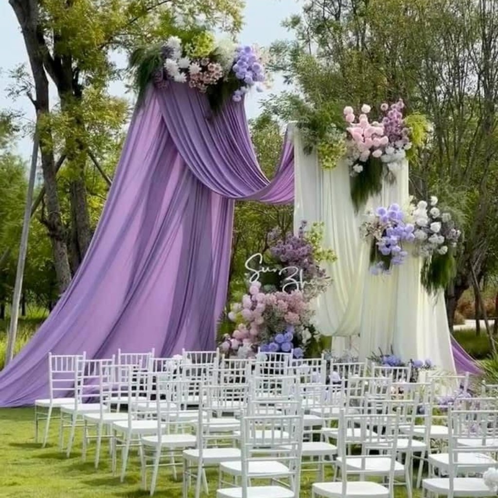  A ceremonial outdoor wedding altar draped in lavender and ivory fabric with cascading floral arrangements in purple, pink, and white. White chairs are set in rows on the lawn, with greenery and trees in the background.