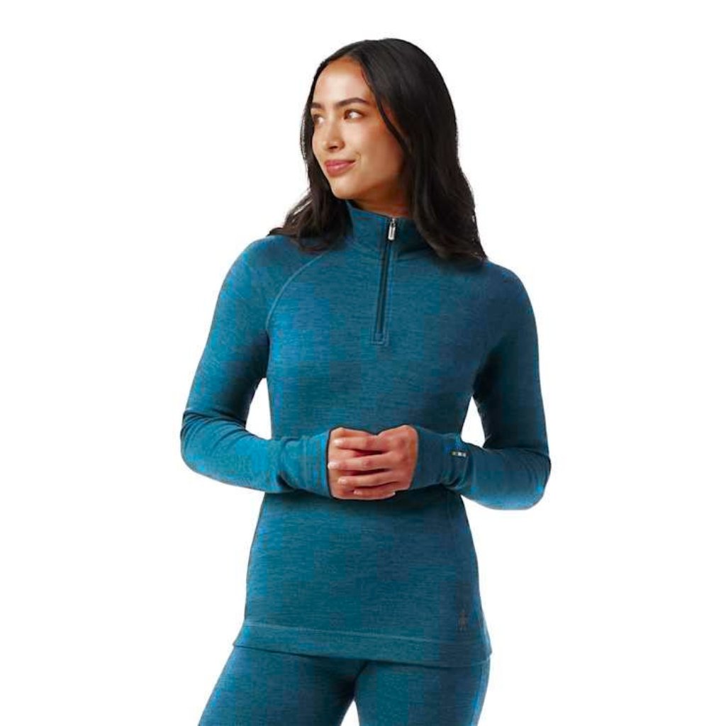 A woman wearing a teal long-sleeve athletic top with a half-zip neck and matching teal leggings, standing with hands clasped.