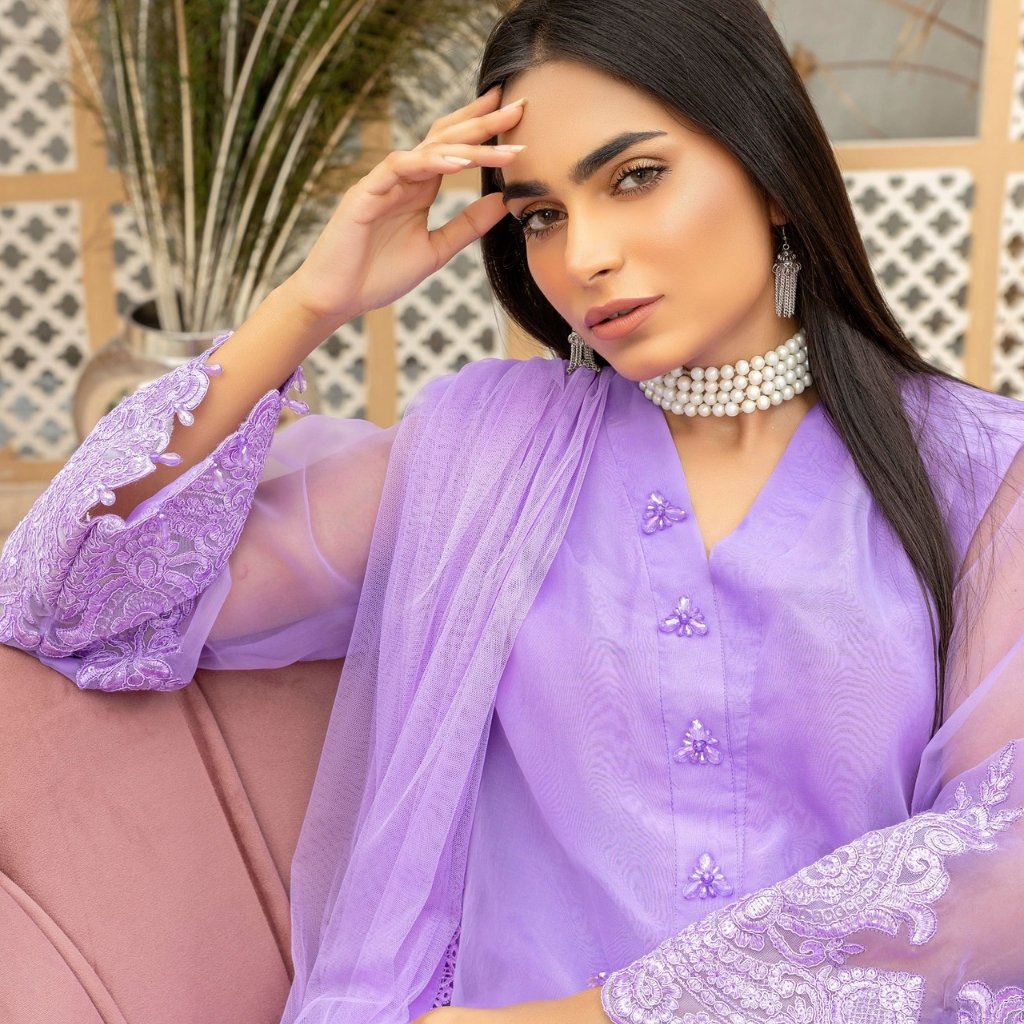 A woman with long dark hair wearing a lavender outfit with lace sleeves and a pearl choker, posing with her hand near her forehead.
