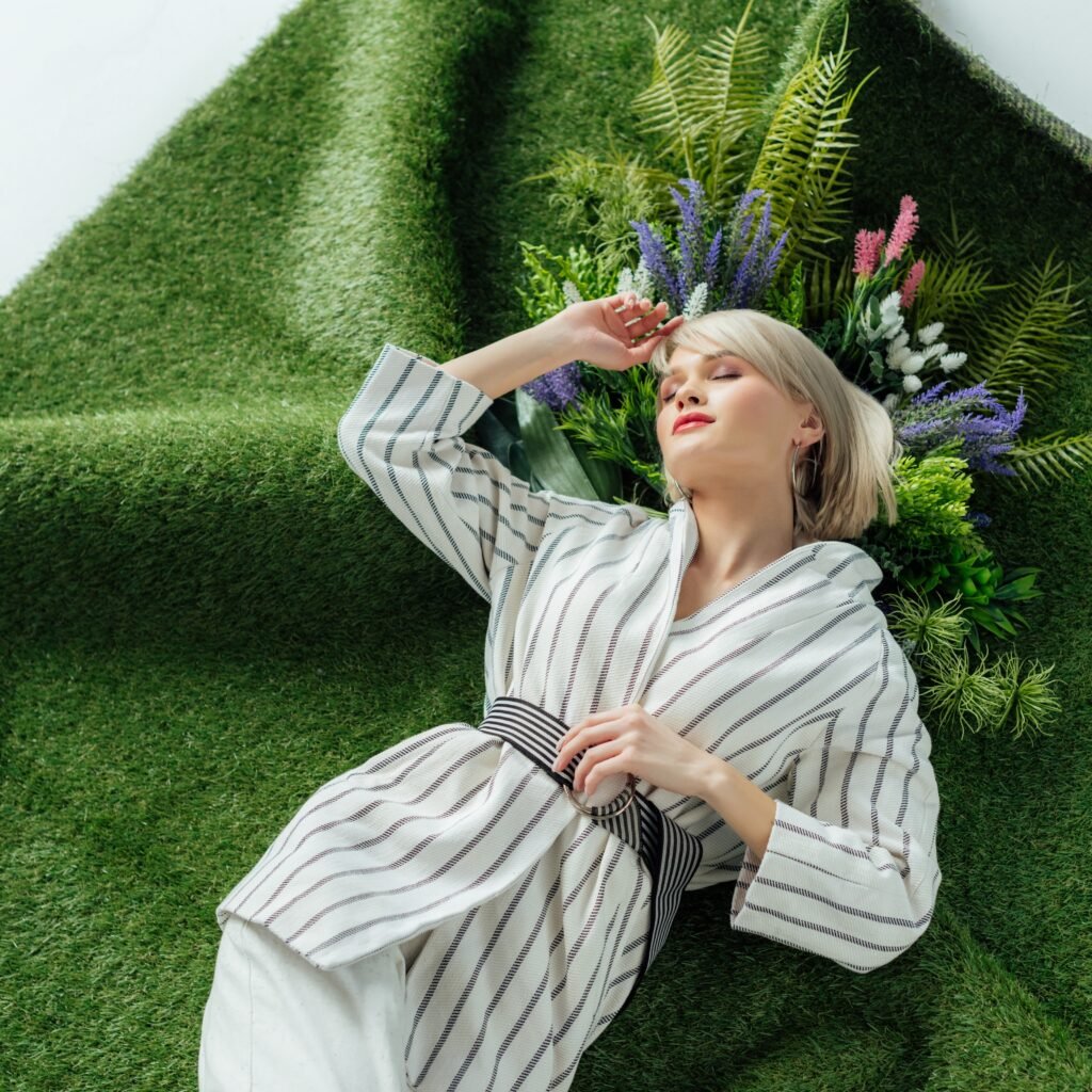 A stylish woman with short blonde hair lounges against a lush, green, grassy backdrop, wearing a light striped robe and a striped belt. She appears relaxed with eyes closed, surrounded by vibrant greenery and colorful flowers.
