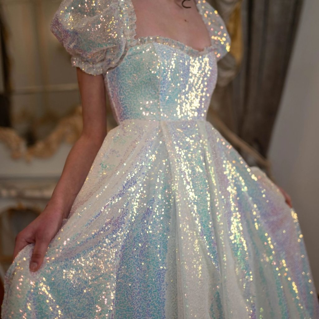 A close-up of a person wearing a sparkling, iridescent ballgown with puff sleeves and a fitted bodice. The dress shimmers with sequins and pastel colors, and the person is holding the skirt to show its fullness.