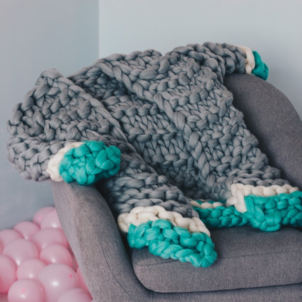 A chunky knitted blanket in soft gray draped over a gray armchair, with teal and white accents. A teal and white knit edge is visible, and pink balloons are in the background.