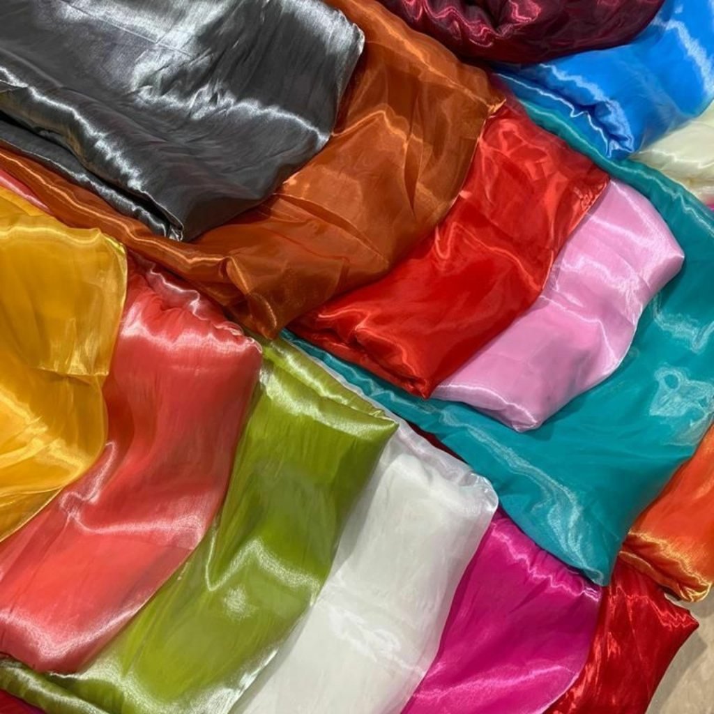  A vibrant assortment of shiny satin fabrics in a rainbow of colors, including yellow, orange, red, pink, green, blue, teal, white, gray, and brown, casually draped and folded together.