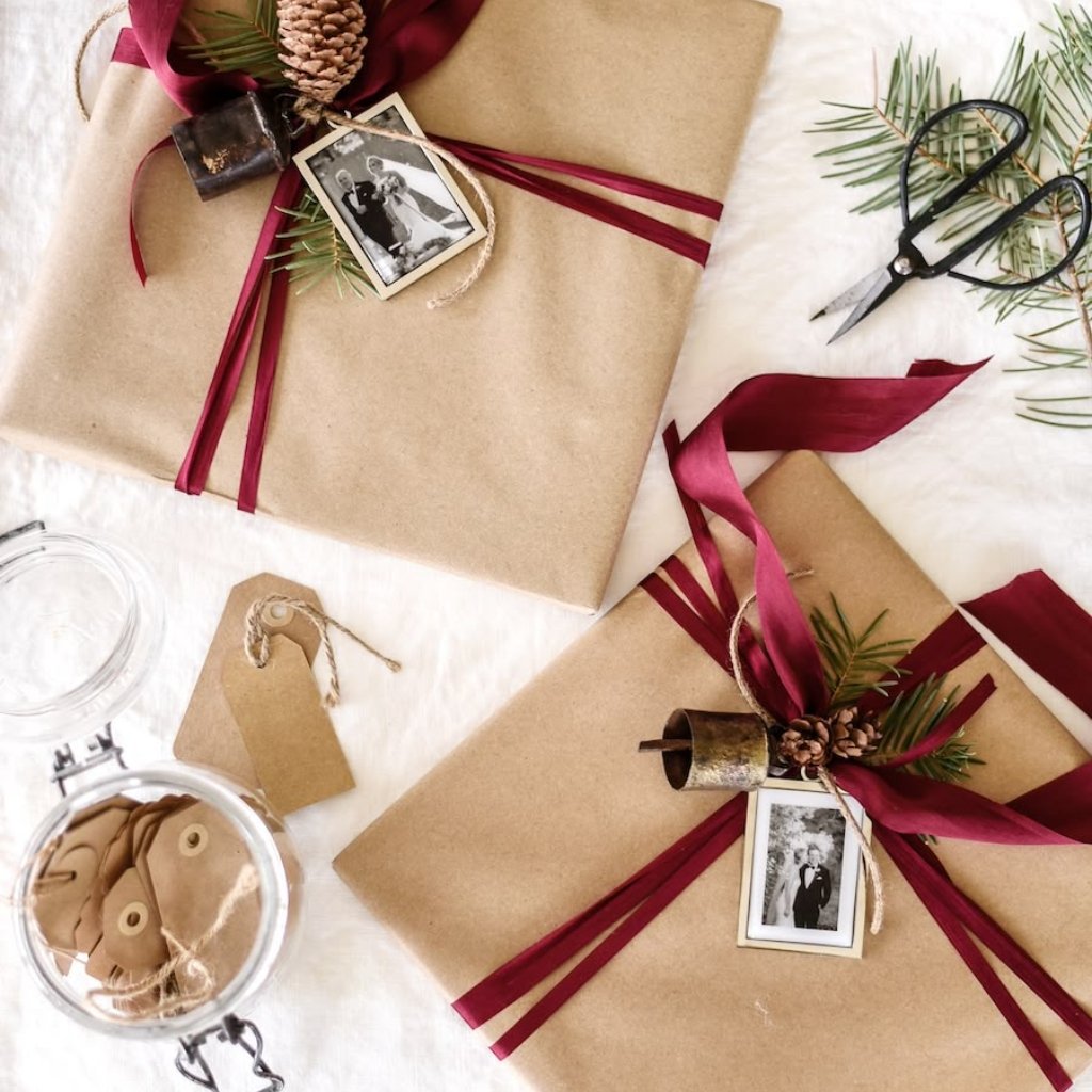 A top-down view of two beautifully wrapped brown paper gifts tied with burgundy ribbons. Each gift has a small vintage black-and-white photo tag attached with twine, pine sprigs, and pinecones. Scissors and evergreen branches sit nearby on a light surface.