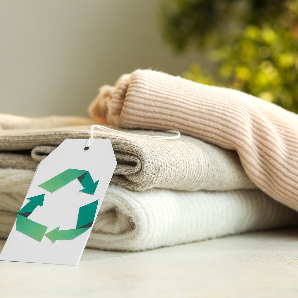 A stack of soft, folded sweaters in neutral tones (beige, cream) with a white clothing tag featuring a green recycling symbol in the foreground.