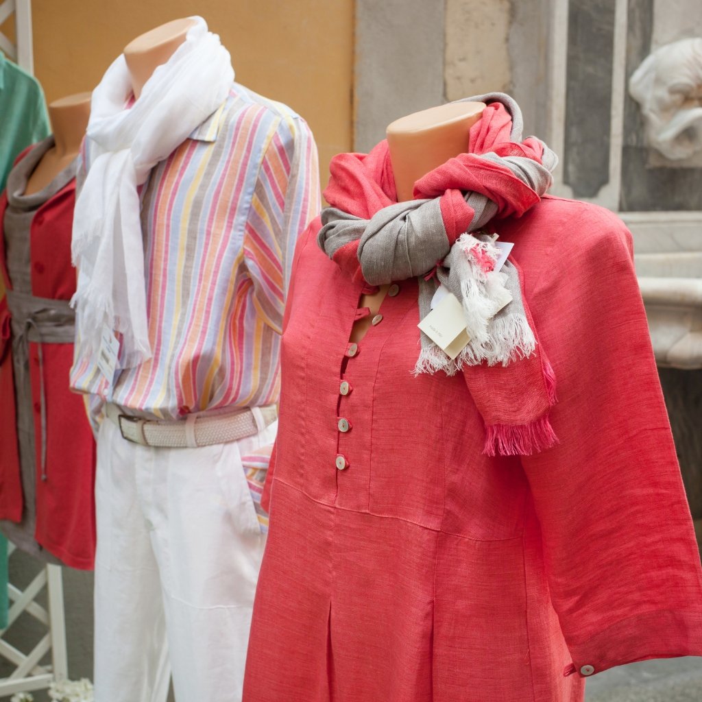 Display of mannequins dressed in chic, comfortable outfits: a coral pink linen dress with a button placket and pleated skirt, paired with a two-tone scarf; a striped pastel shirt with white pants and a light belt; and other soft, casual ensembles in reds and neutrals.