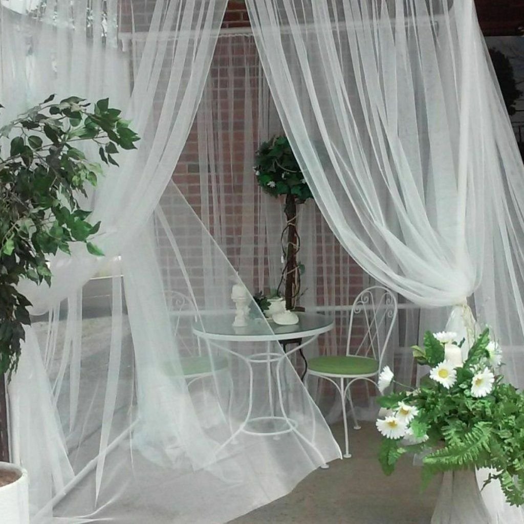 White Organza Fabric: Elegant Style for Dresses and Interiors 26 A delicate white outdoor seating area draped with sheer white fabric curtains, a small round glass table with two white wrought-iron chairs, green plants and a floral arrangement with white daisies in the foreground.