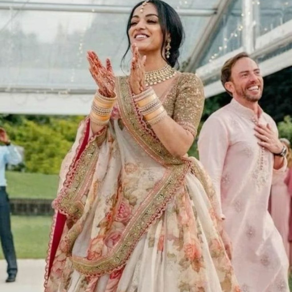  A joyful woman wearing a pale floral lehenga with gold embroidery and matching jewelry claps her hands at an outdoor celebration, while a man in a light pink traditional kurta smiles in the background.