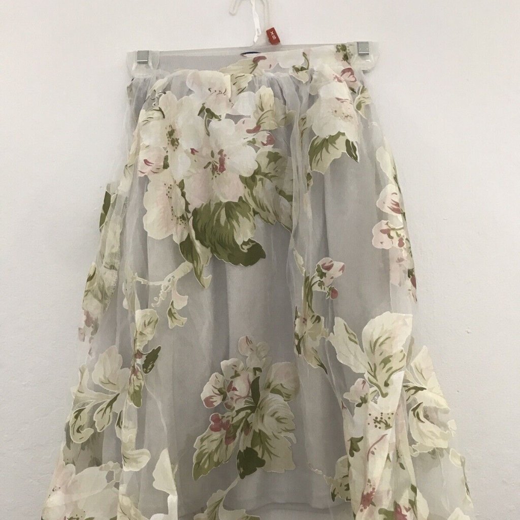  A light, sheer floral skirt hanging on a white wall. The fabric is translucent with large cream and pale pink flowers in green leaves, draping in soft layers from a hanger.