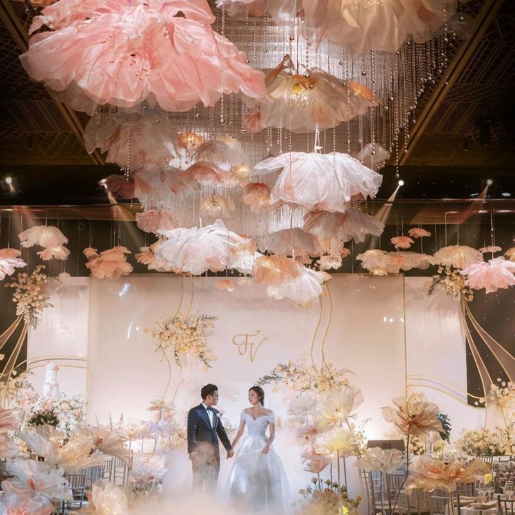 A bride and groom stand hand in hand in a pastel wedding venue filled with large, delicate pink and white paper-like floral decorations hanging from the ceiling; soft mist covers the floor as guests' chairs surround an elegant, ivory backdrop.
