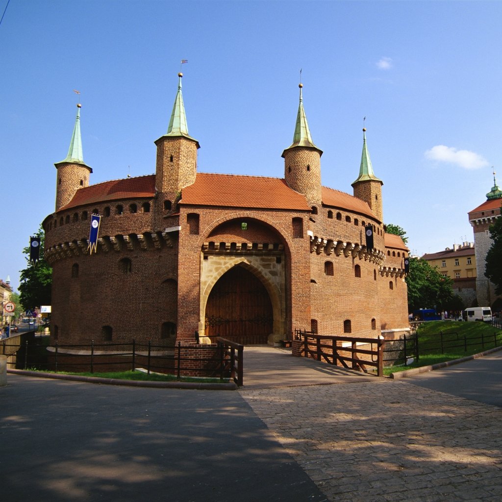 A historic brick fortress with rounded towers and green conical roofs sits at a cobblestone street entrance. The structure has arched gateways, small windows, and decorative battlements, set against a clear blue sky.