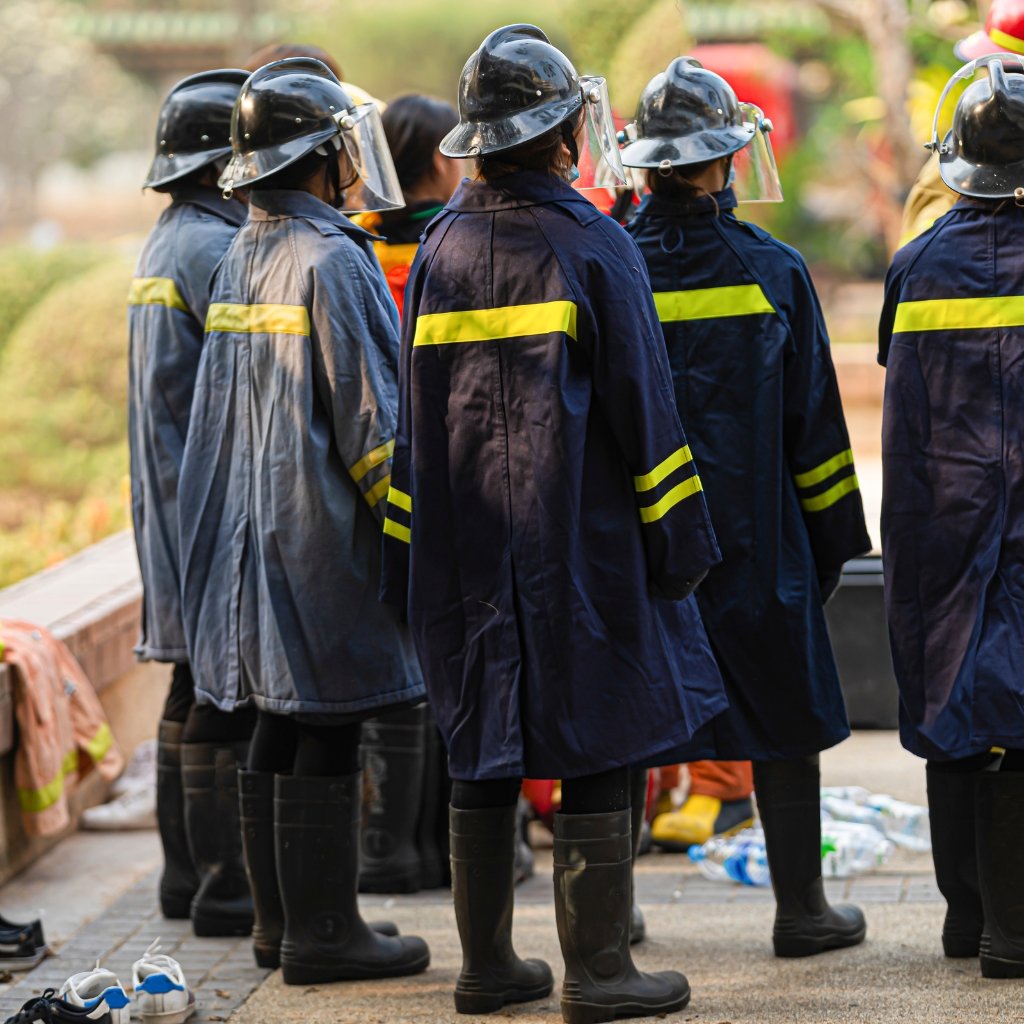 Group of firefighters wearing dark blue uniforms with reflective yellow stripes and helmets, standing outdoors on a paved area; scattered footwear and gear on the ground nearby, blurred background.