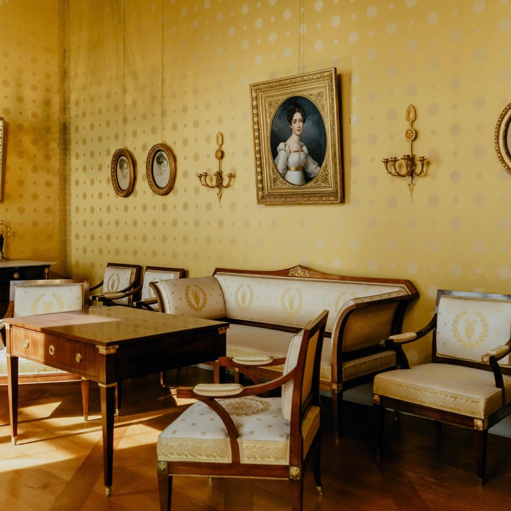 A vintage-inspired living room with yellow patterned walls, gold-framed portraits, and cream-upholstered seating. A wooden table sits in the center, surrounded by matching chairs and a sofa, all trimmed with gold accents. Soft sunlight streams across the polished wooden floor.