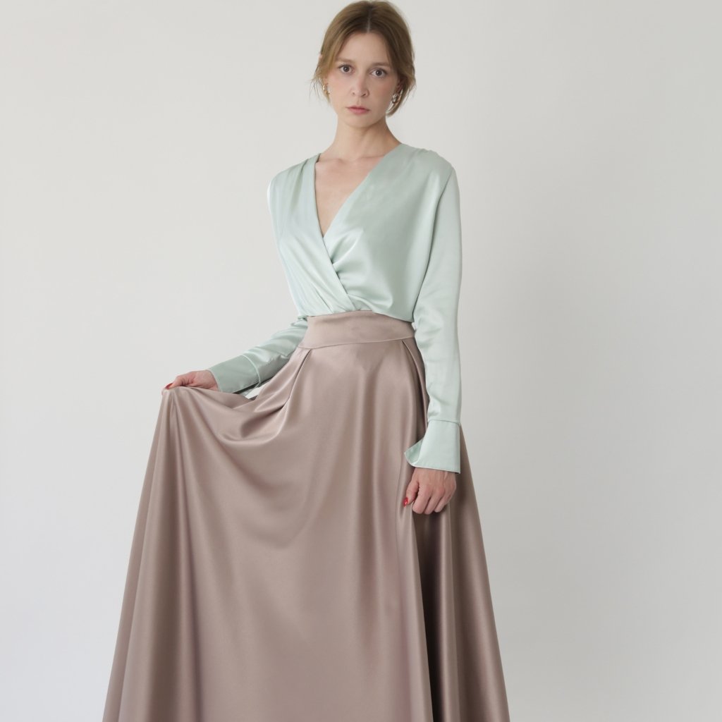 A fashion model sits gracefully against a light gray wall, wearing a soft mint-green satin wrap-top with long sleeves and a high-waisted taupe satin skirt. The fabric has a subtle sheen and fluid drape.