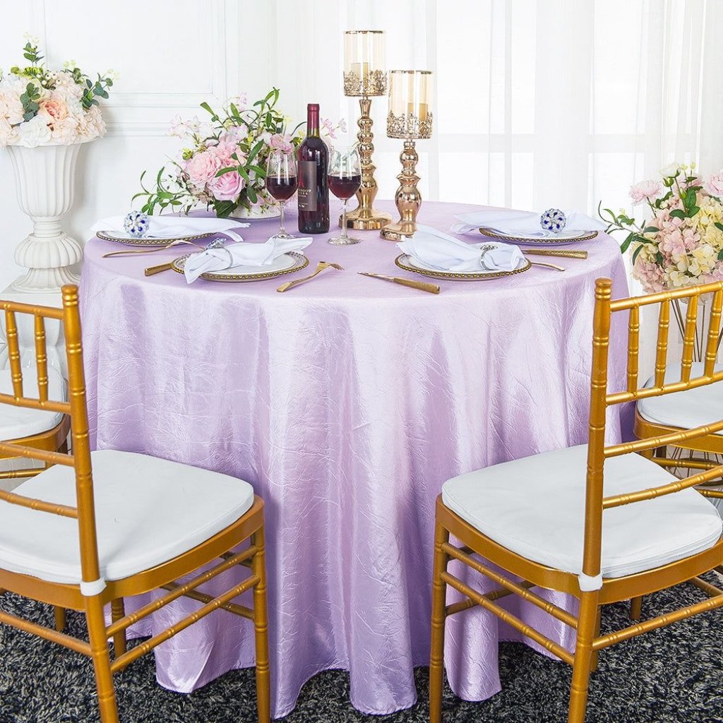 A round banquet table draped in a lavender tablecloth with gold chairs; elegant place settings, wine bottles, candles, and floral arrangements.

