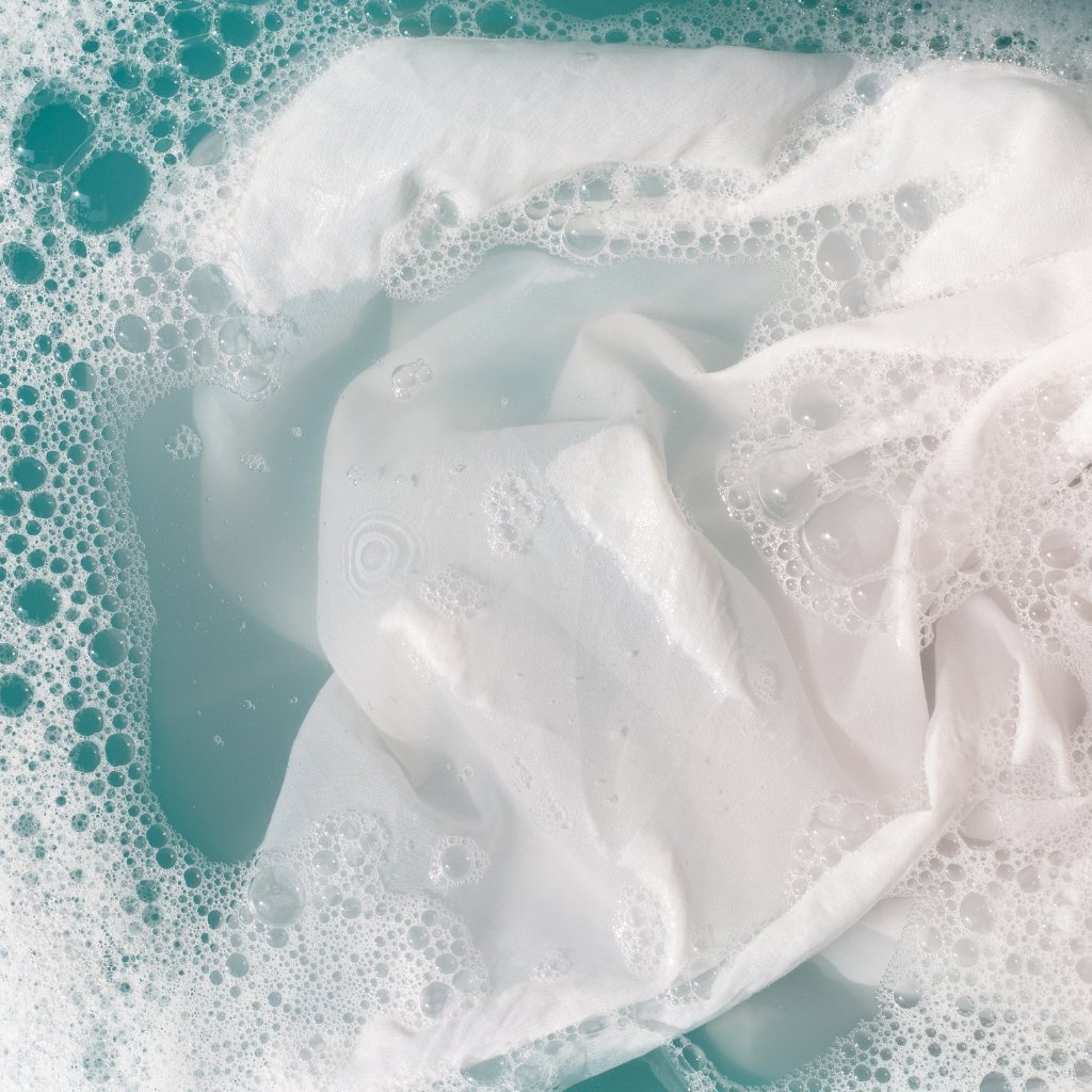 A close-up of white fabric being washed in soapy water with lots of bubbles around it.
