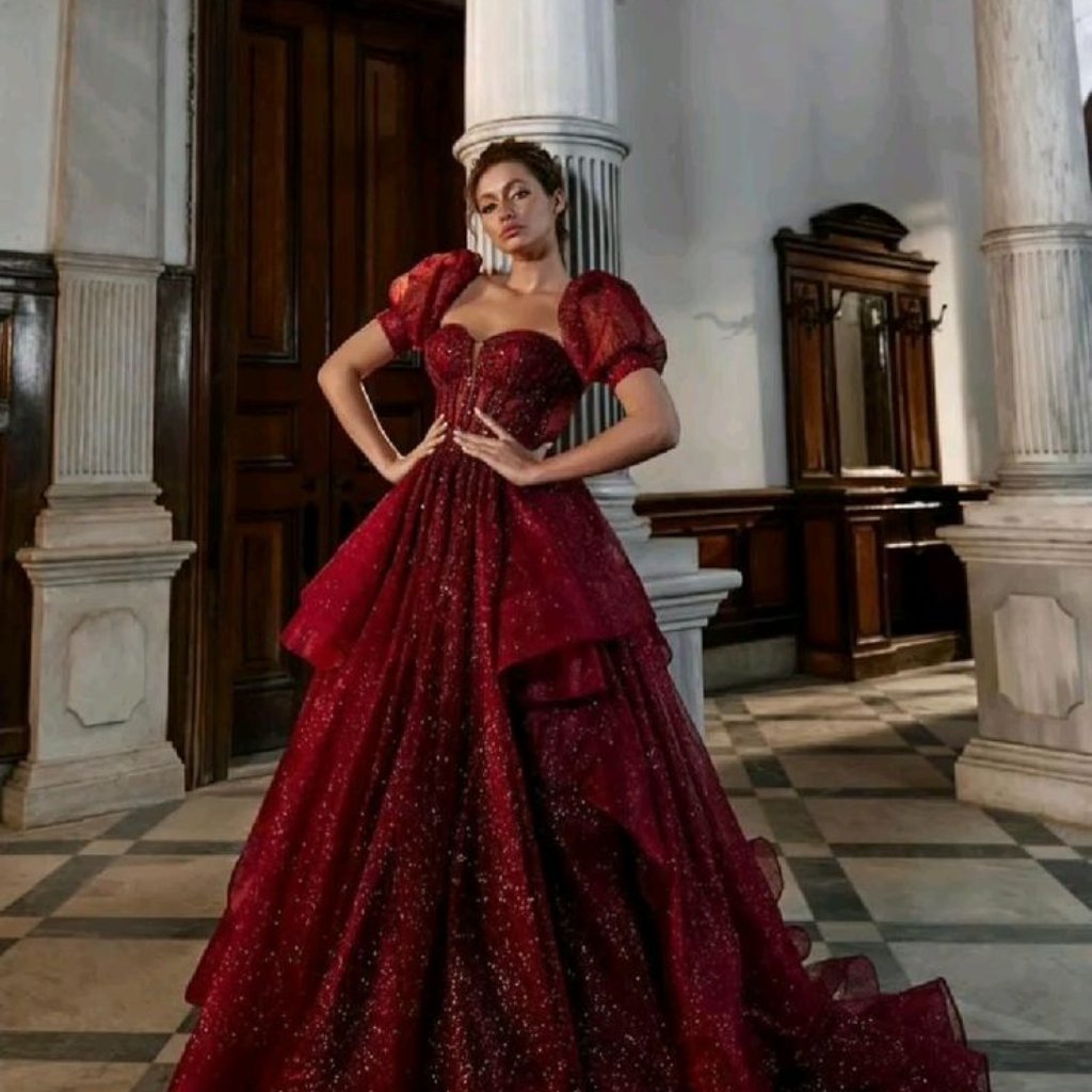 A woman stands in a grand indoor hall wearing a dramatic, sparkling red ballgown with puff sleeves and a voluminous layered skirt. She poses with hands on her hips beside marble columns and antique furniture.

