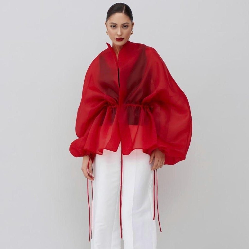 A woman wearing a bold red sheer cape-style top with voluminous sleeves and a gathered waist, paired with white wide-leg trousers against a light gray background.