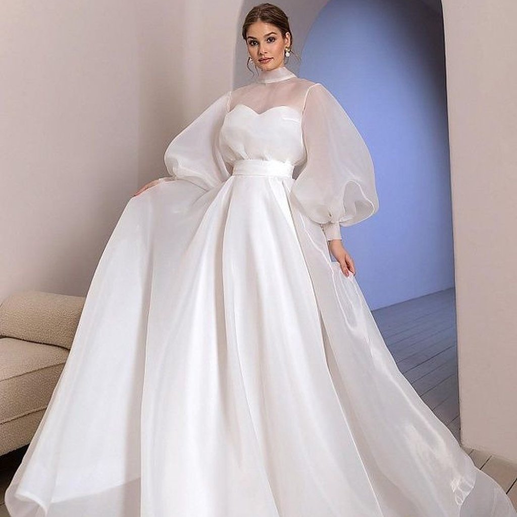 White Organza Fabric: Elegant Style for Dresses and Interiors 20 A bride wearing a white wedding gown with a fitted heart-shaped bodice, sheer high neckline, and dramatic, billowing puff sleeves. The dress has a defined waist and a full A-line skirt that sweeps the floor, with a soft train visible as she stands in a minimalist interior.