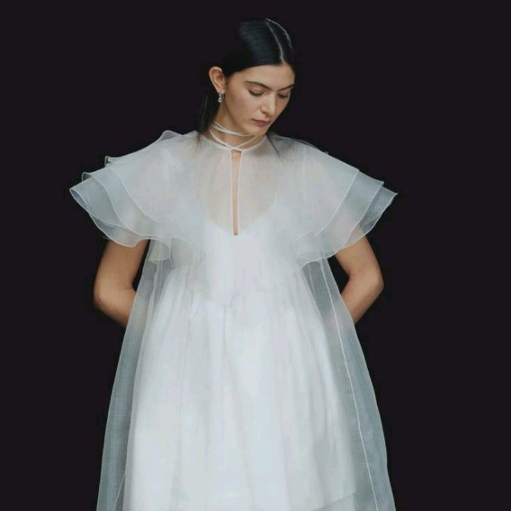 White Organza Fabric: Elegant Style for Dresses and Interiors 32 A woman wearing a delicate white sheer cape over a white dress, standing against a dark background with her head slightly bowed.