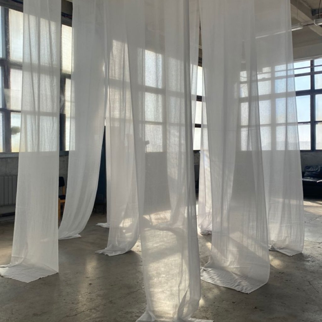 White Organza Fabric: Elegant Style for Dresses and Interiors 24 Long, flowing white sheer curtains or drapes hanging from the ceiling in a bright, industrial-style room with large windows. The fabric pools slightly on the concrete floor, creating soft shadows and light patterns.
