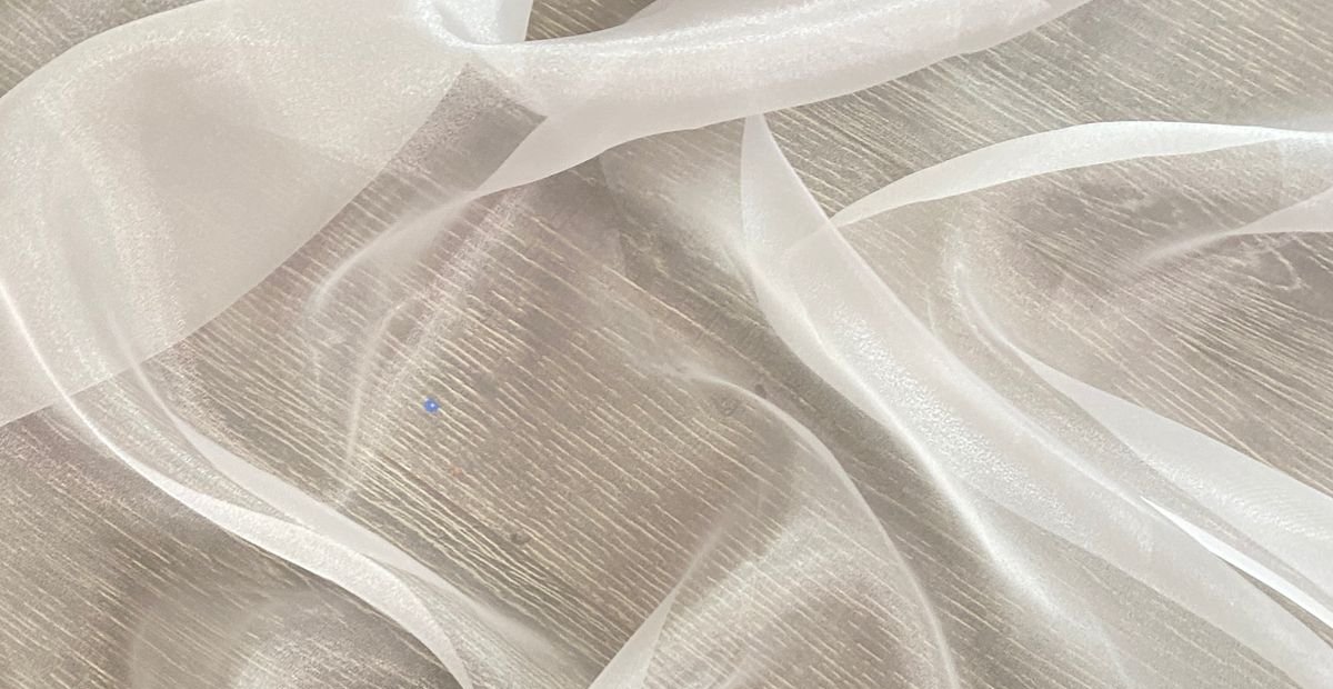 A close-up of sheer, white ribbon-like fabric laid on a light wooden surface, with gentle folds and translucent texture. White Organza Fabric