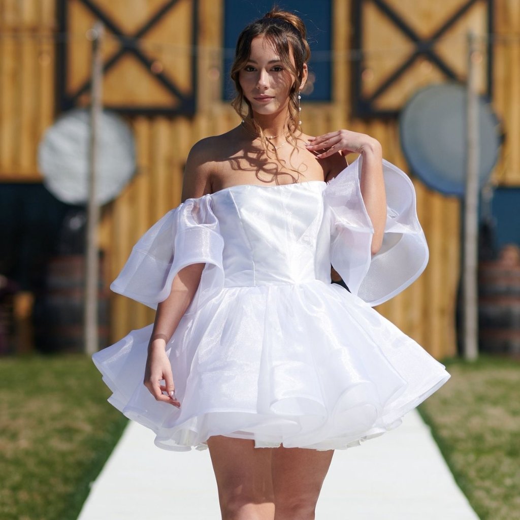 White Organza Fabric: Elegant Style for Dresses and Interiors 22 A young woman wearing a short, off-the-shoulder white dress with voluminous sheer sleeves stands on a runway outdoors. She looks at the camera with a soft expression, and the background shows a wooden building and a sunny day.