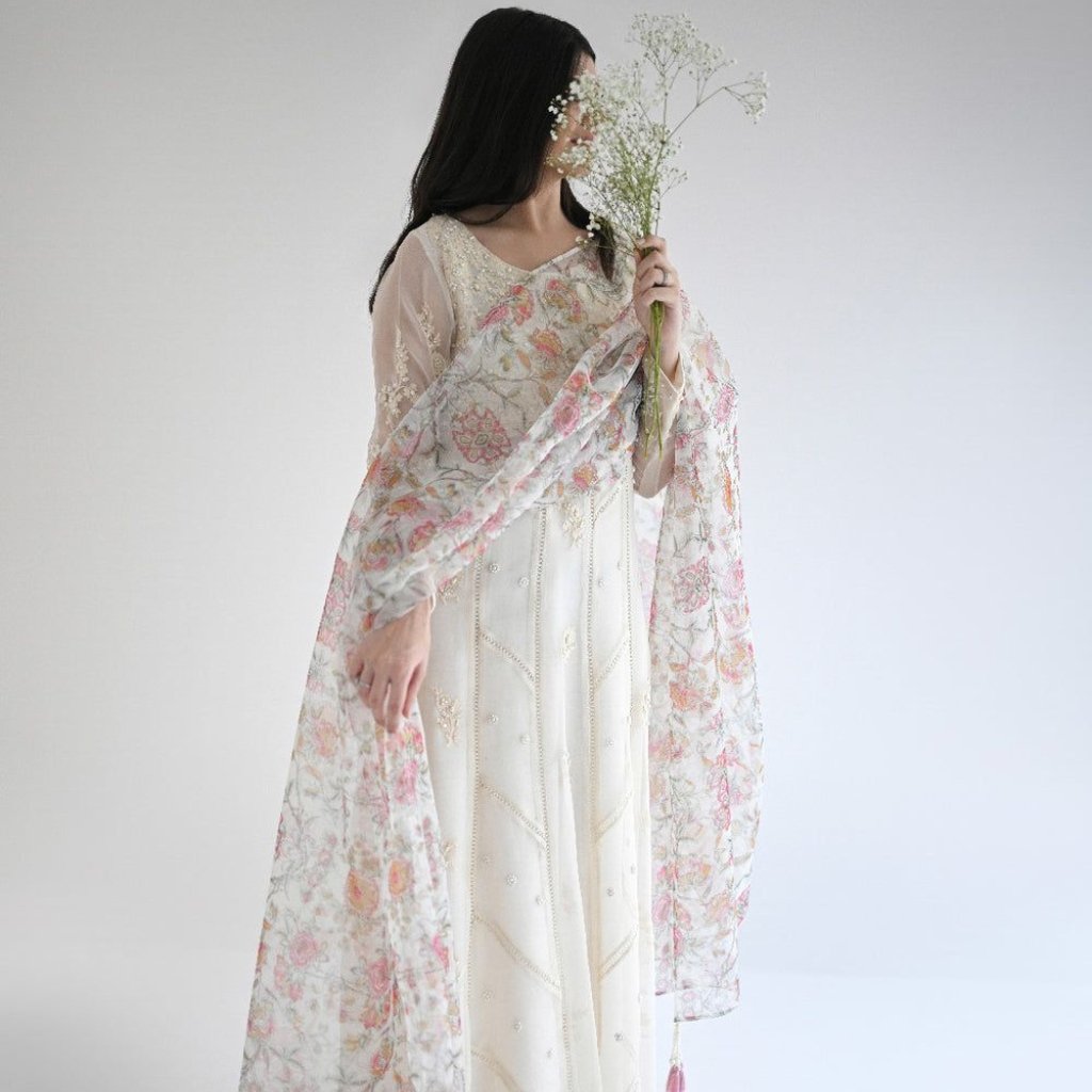 White Organza Fabric: Elegant Style for Dresses and Interiors 23 A woman wearing an elegant light-colored wedding or formal dress with delicate floral embroidery, holding a bouquet of small white flowers. She drapes a sheer, floral scarf over her shoulders and partially covers her face with the flowers.