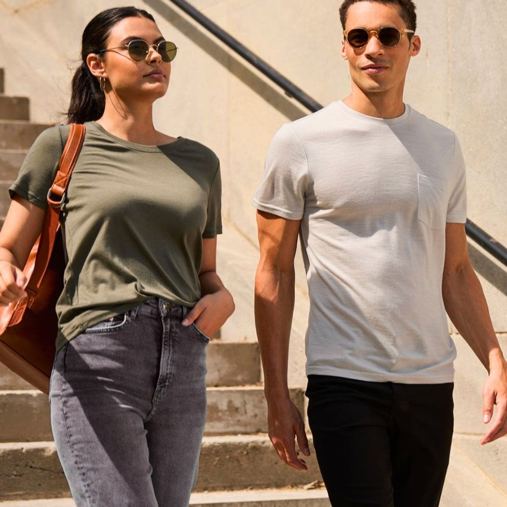 A stylish man and woman walk down sunlit outdoor steps, both wearing casual t-shirts and sunglasses; the woman carries a brown shoulder bag and wears grey jeans, while the man wears black pants.