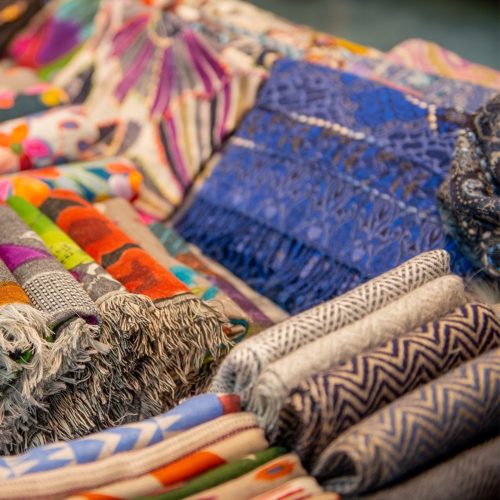 Close-up view of rolled and folded wool blankets in various colors and patterns, including camouflage-inspired earth tones, bright stripes, and intricate weaves, with a fringed edge on one roll.