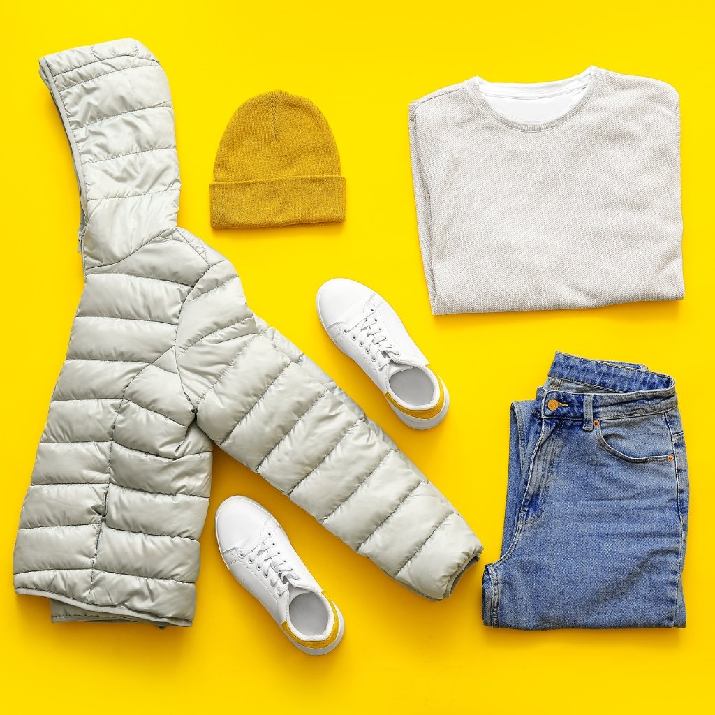 A flat lay of casual winter clothing on a bright yellow background: a light gray puffer jacket, a mustard knit beanie, a folded light gray knit top, blue denim jeans, and a pair of white sneakers.