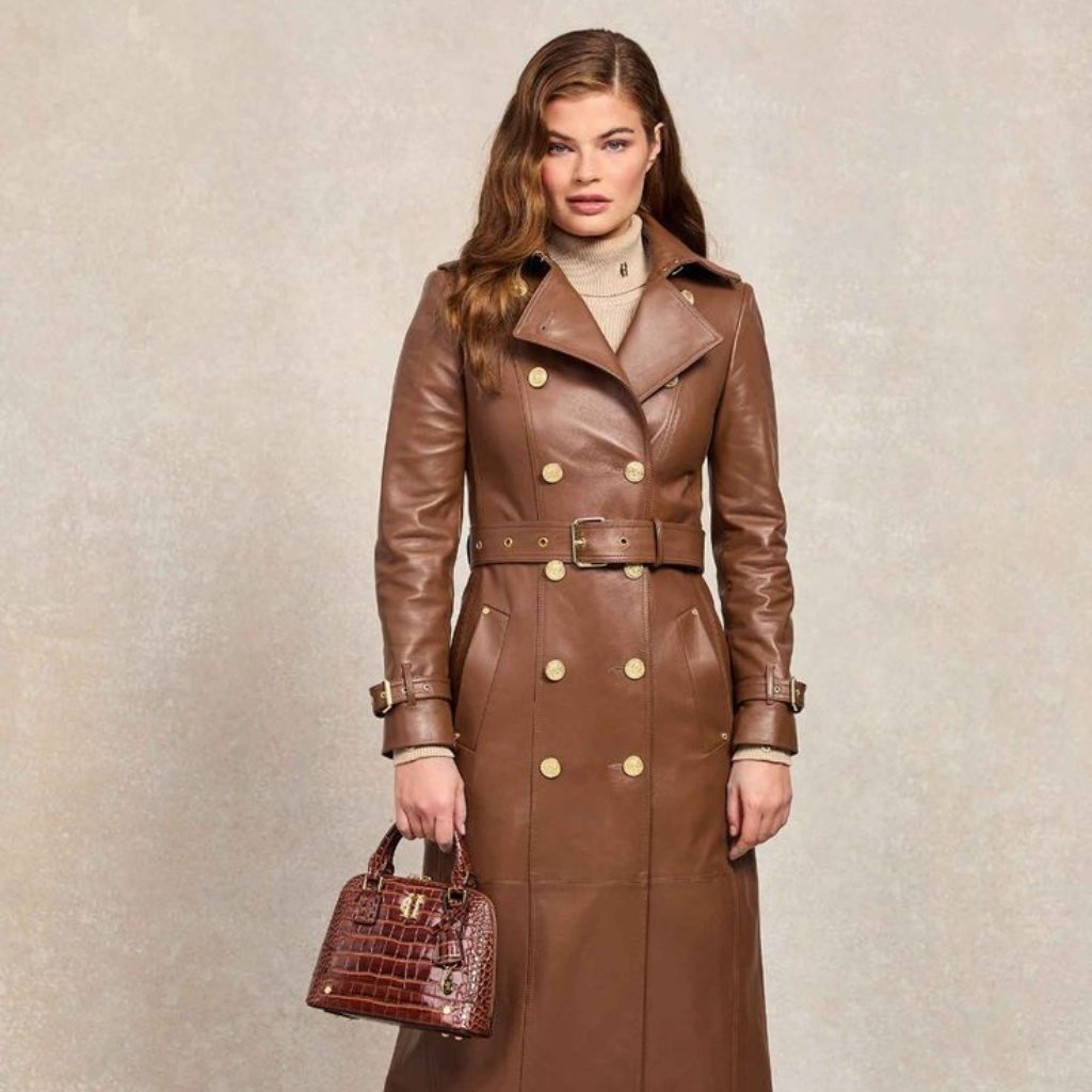 A woman wearing a long brown leather trench coat with a double-breasted front and wide lapels. She holds a small brown crocodile-embossed handbag and wears a beige turtleneck underneath. The coat features a belt at the waist and buckle straps at the cuffs.