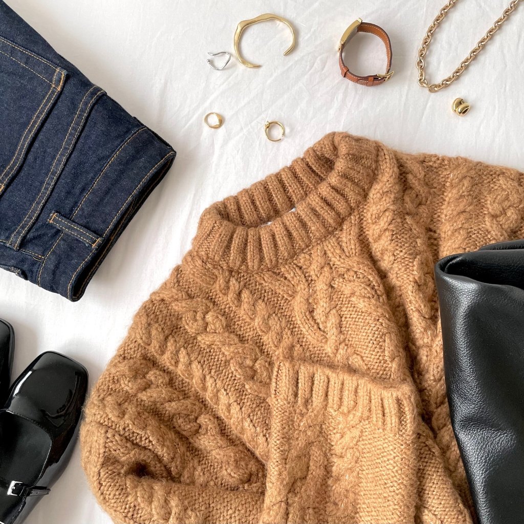 A cozy flat lay featuring a chunky camel knit sweater, a pair of dark blue denim jeans, black heeled shoes, and a black leather bag. Jewelry pieces (rings, bracelets) and a chain necklace are scattered around on a white background.
