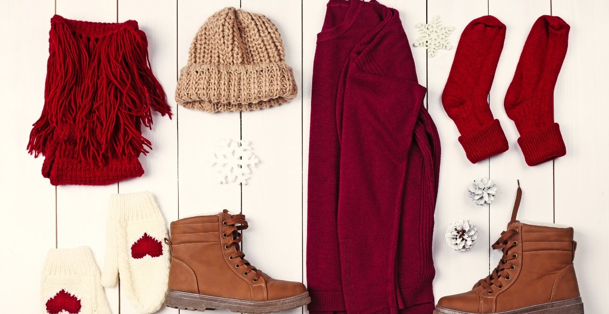 Guide to Wool Outfits: Warm, Stylish & Sustainable Flat lay of winter clothing and accessories: red scarf, beige knit hat, maroon sweater or coat, matching maroon socks, white mittens with red hearts, tan lace-up boots, and decorative pinecones on a white wooden background. Wool Outfits