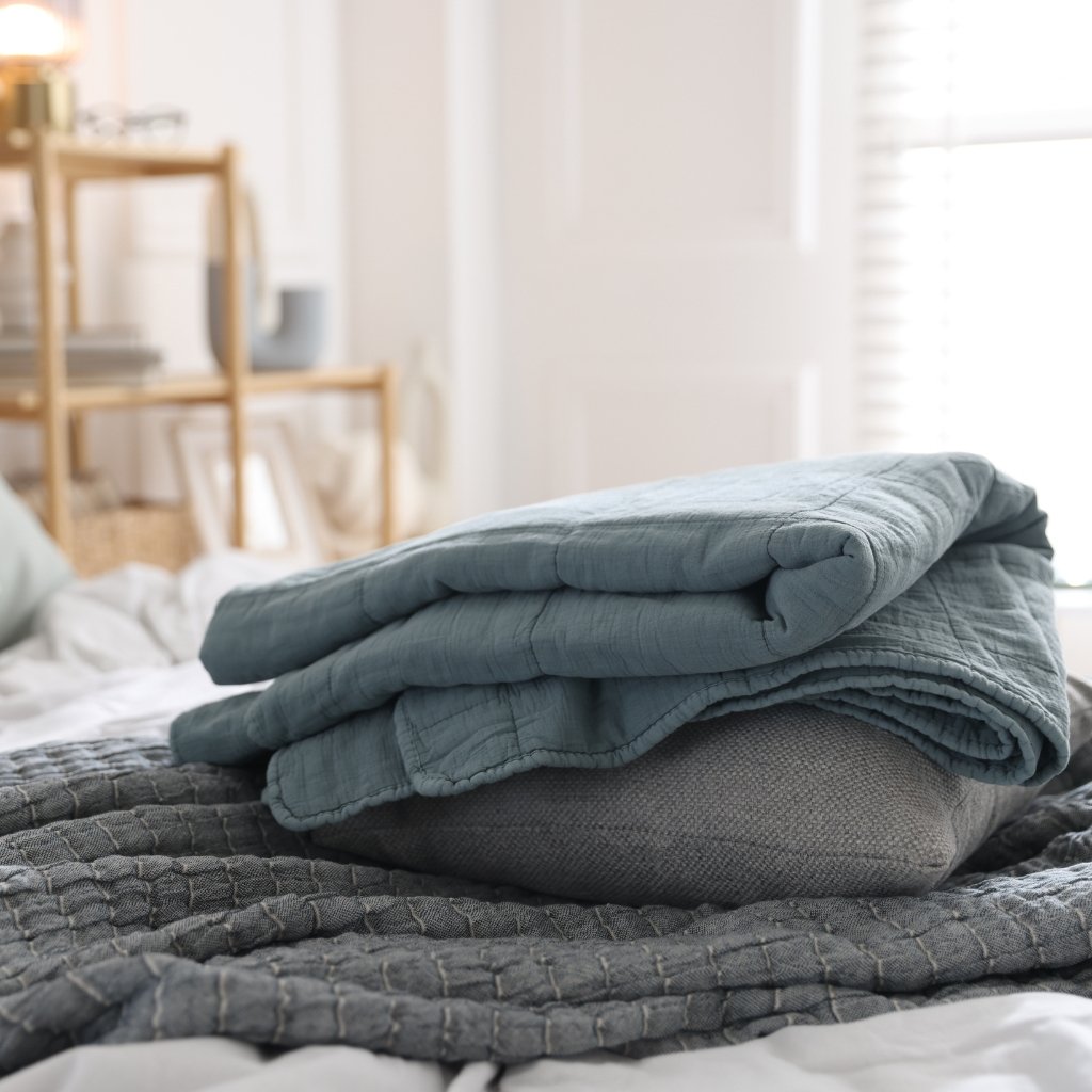 Why Seersucker Cotton Fabric Is Perfect for Summer Outfits 15 Folded blue quilt resting on a gray cushion atop a textured blanket in a softly lit bedroom.