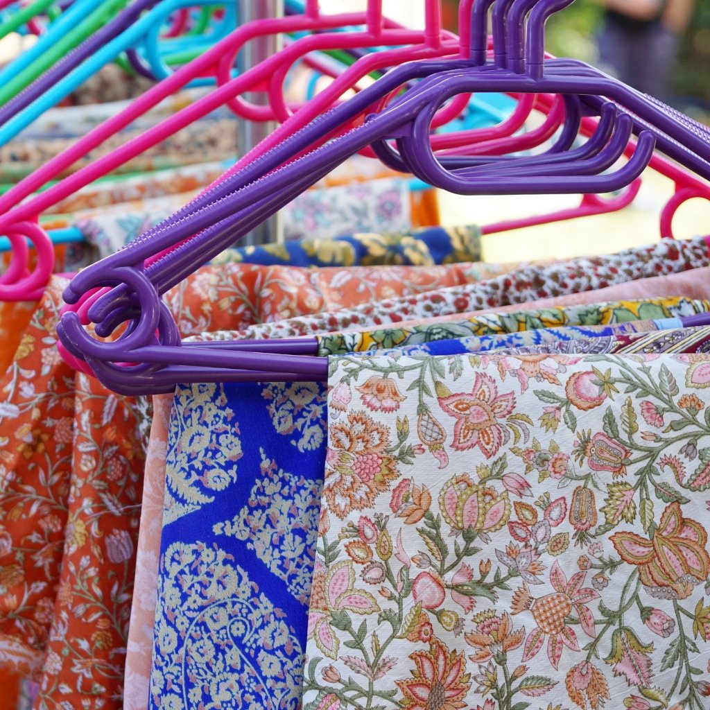 A close-up of colorful plastic clothing hangers (purple, pink, blue, and green) hanging on a rack with patterned fabric clothes in bright floral designs.